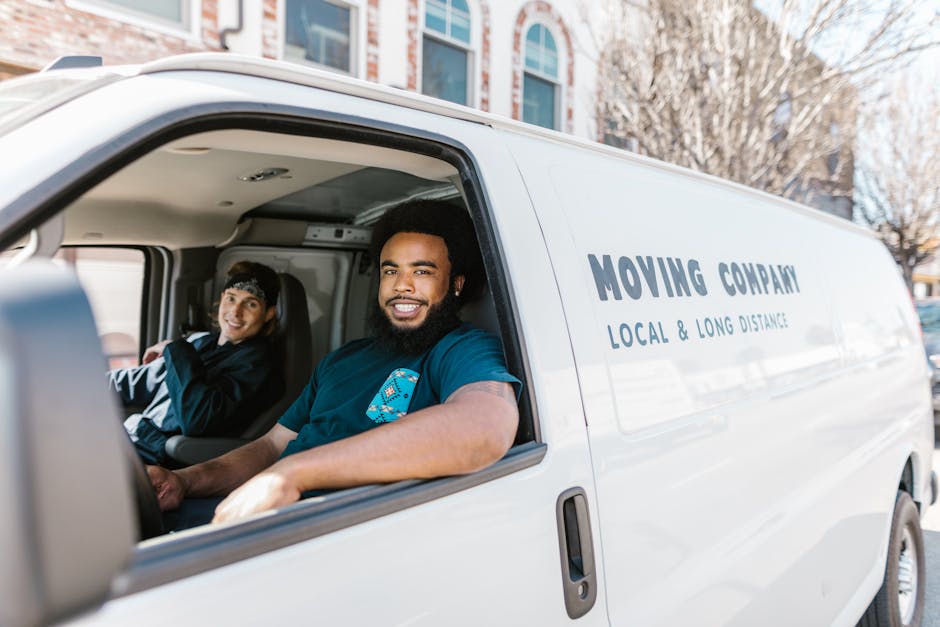 A man wearing a white cap and a dark vest over a blue long-sleeved shirt stands outside a white moving van, which is parked on a paved area near a modern building with large glass windows and metallic