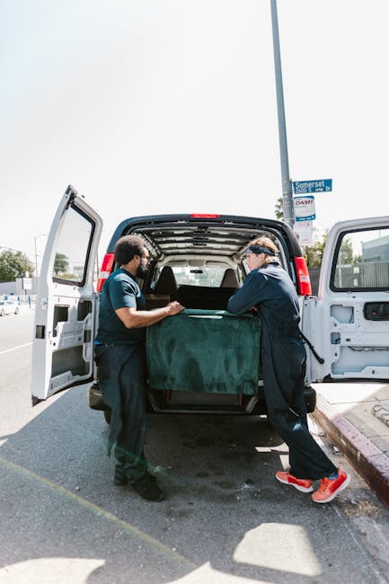 A man wearing a headband and dark clothing is outside on a street, carrying a large, wrapped piece of furniture with both hands. The furniture is covered in green and black protective materials, secur
